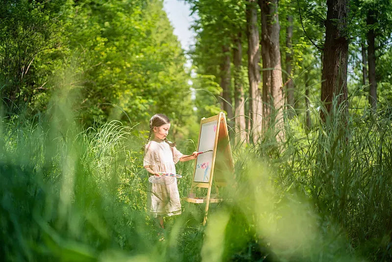 A young girl painting on an easel in a lush green forest. A visual metaphor for being entirely present in the physical world, illustrating the somatic focus that Jambhala grounding jewelry helps adults reclaim.