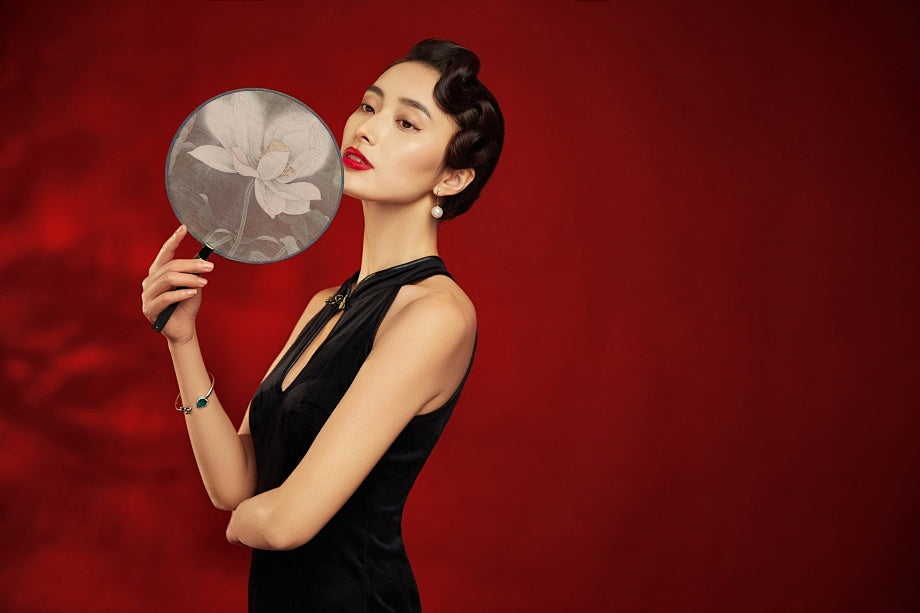 An elegant woman holding a traditional round fan against a deep red background. A visual representation of modern eastern aesthetic, reflecting the timeless physical connection between human touch and authentic natural stone jewelry.