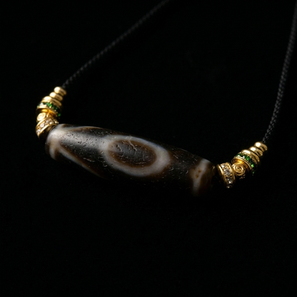 Close-up of the Calm Witness Dzi Agate grounding necklace, featuring a striking 46.5mm weathered cylindrical stone with an eye motif and 18K gold pavé spacers on a dark background.