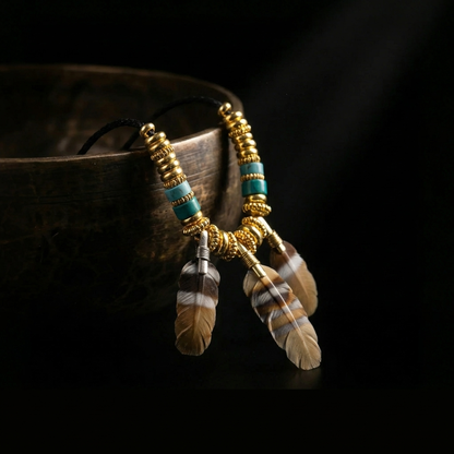 The Quiet Feather grounding necklace resting against an antique brass bowl, highlighting the deeply grooved agate feathers and turquoise accents in dramatic lighting.
