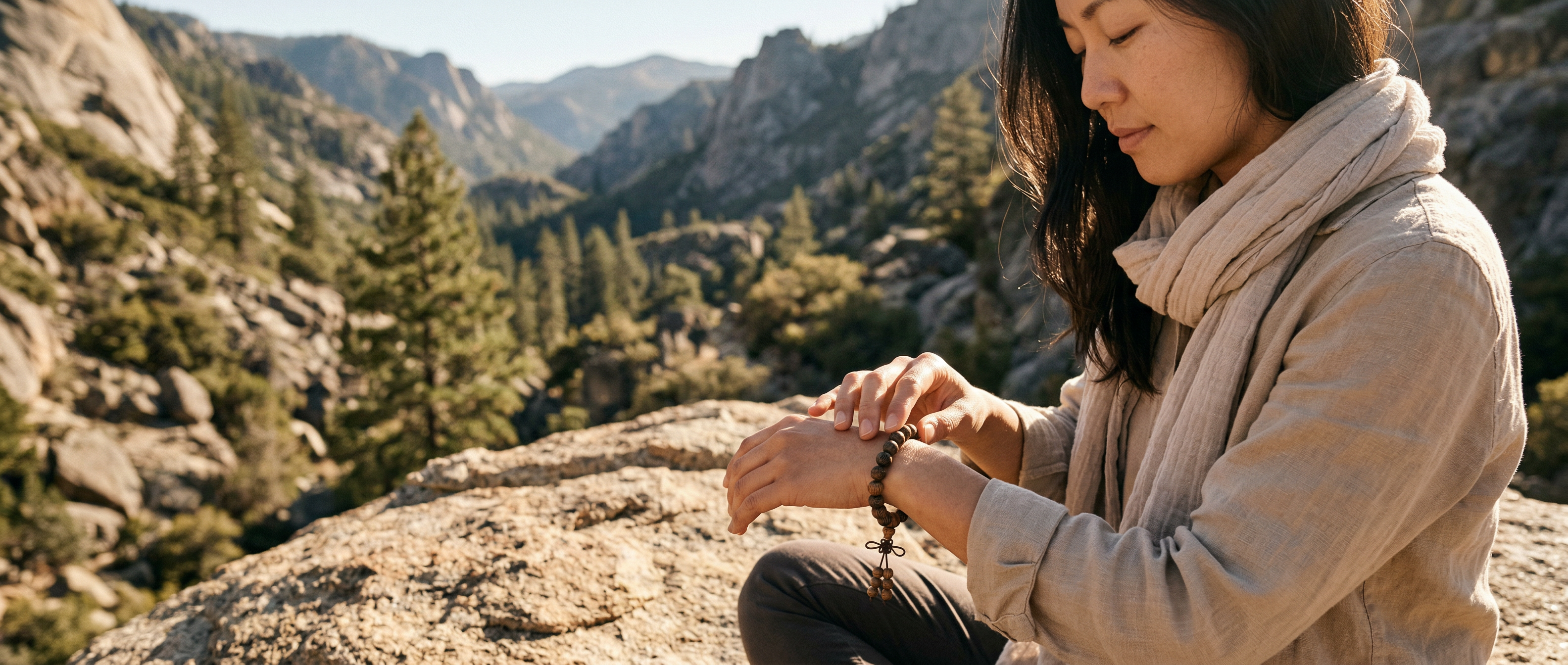 Woman in a neutral linen outfit resting on a rocky mountain outcrop, tracing the beads of her dark wood grounding bracelet. Showcasing effortless modern eastern aesthetic, the tactile wooden beads serve as a quiet physical anchor to cut through mental noise.