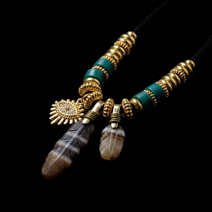 Close-up of the grounding charm necklace, featuring two hand-carved banded agate feathers, a golden sentinel eye charm with crystal accents, cylindrical turquoise beads, and 18K gold-plated ribbed spacers.
